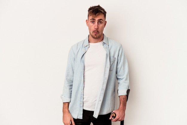 Young caucasian man holding crutch isolated on white background shrugs shoulders and open eyes confused.