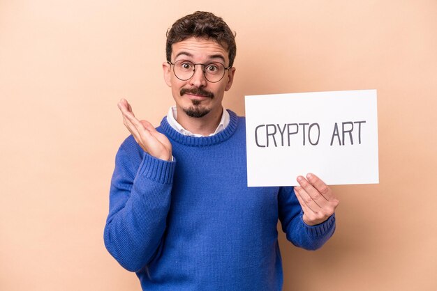 Young caucasian man holding a crypto art placard isolated on beige background surprised and shocked.