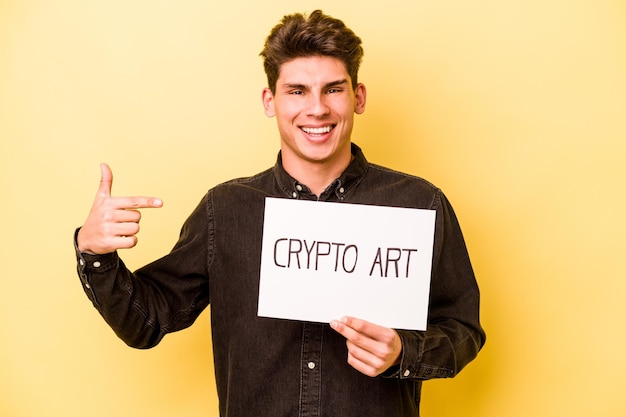 Young caucasian man holding crypto art placard isolated on yellow background person pointing by hand to a shirt copy space proud and confident