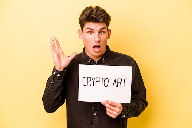 Young caucasian man holding crypto art placard isolated on yellow background surprised and shocked