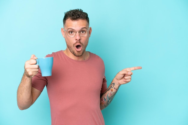 Young caucasian man holding cup of coffee isolated on blue background surprised and pointing side