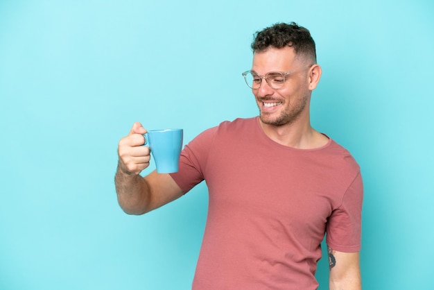 Молодой кавказский мужчина держит чашку кофе на синем фоне со счастливым выражением лица