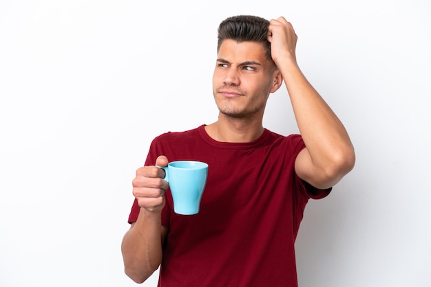 흰색 배경에 격리된 커피 한 잔을 들고 있는 백인 청년은 의심을 품고 얼굴 표정을 혼란스럽게 합니다.