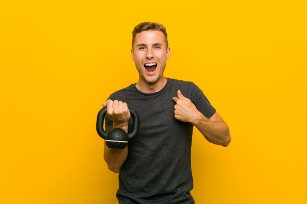 Il giovane uomo caucasico che tiene un dumbbell ha sorpreso indicando se stesso, sorridendo ampiamente.