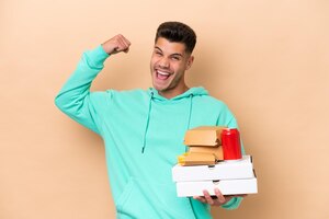 Young caucasian man holding fast food isolated on beige background celebrating a victory