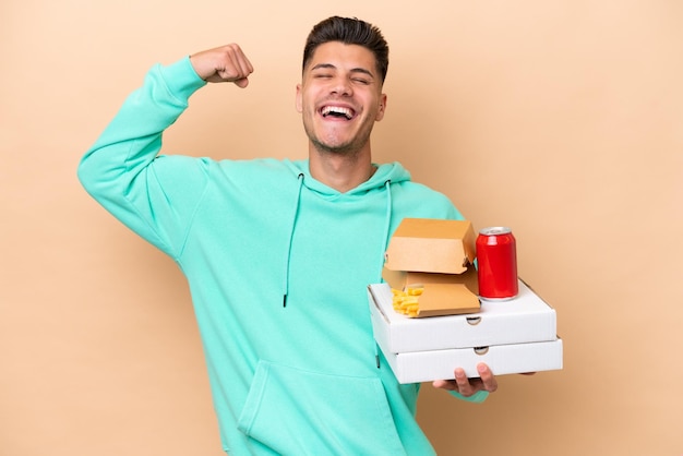 Young caucasian man holding fast food isolated on beige background doing strong gesture