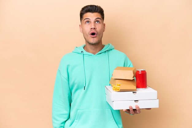 Young caucasian man holding fast food isolated on beige background looking up and with surprised expression