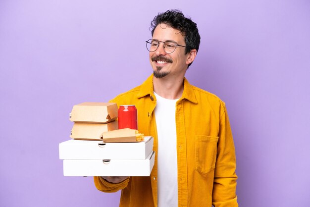 Young caucasian man holding fast food isolated on purple background looking to the side and smiling