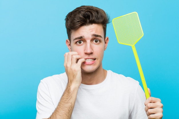 Young caucasian man holding a fly swatter biting fingernails, nervous and very anxious.