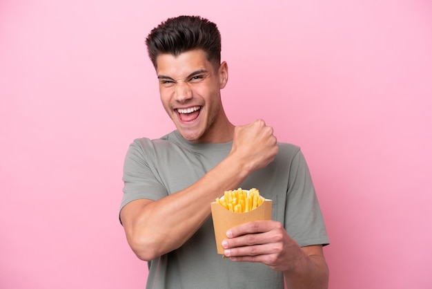 Young caucasian man holding fried chips isolated on pink background celebrating a victory