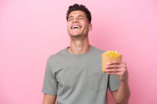 Young caucasian man holding fried chips isolated on pink background laughing