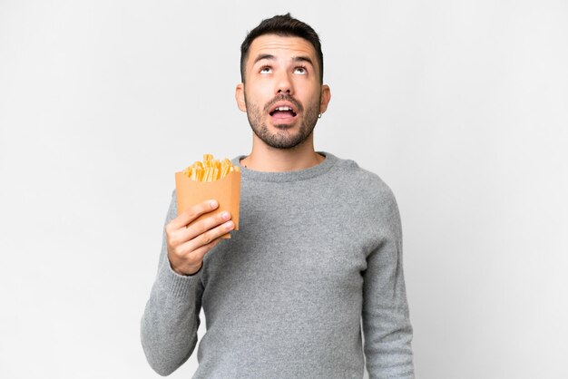 Young caucasian man holding fried chips over isolated white background looking up and with surprised expression