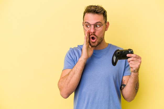 Young caucasian man holding a game controller isolated on yellow background is saying a secret hot braking news and looking aside