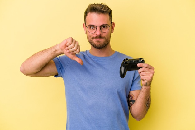 Young caucasian man holding a game controller isolated on yellow background showing a dislike gesture, thumbs down. Disagreement concept.