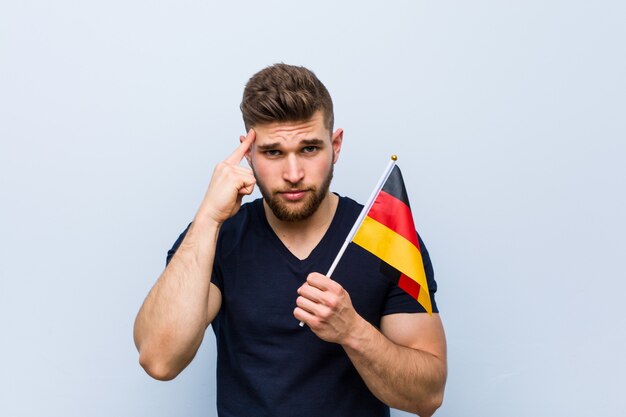 Молодой кавказский мужчина держит флаг Германии, указывая пальцем на висок, думая, сосредоточился на задаче.