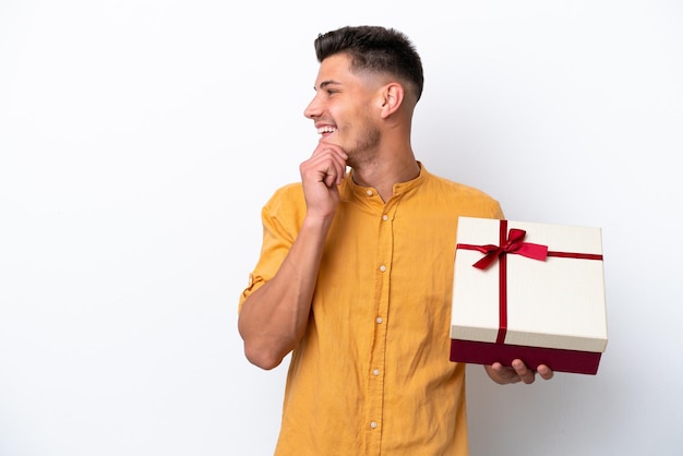 Giovane uomo caucasico in possesso di un regalo isolato su sfondo bianco pensando un'idea e guardando il lato