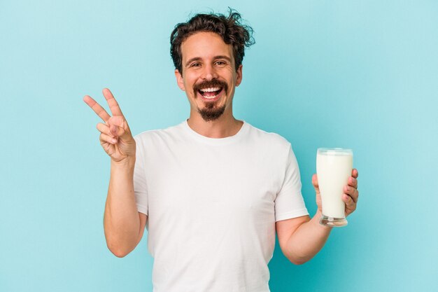青の背景に分離されたミルクのガラスを持っている若い白人男性は、指で平和のシンボルを示して楽しくてのんきです。