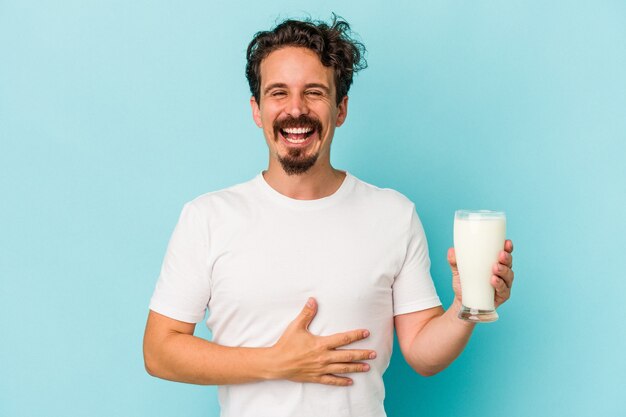 Young caucasian man holding a glass of milk isolated on blue background laughing and having fun.