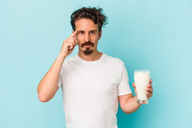 Young caucasian man holding a glass of milk isolated on blue background pointing temple with finger, thinking, focused on a task.