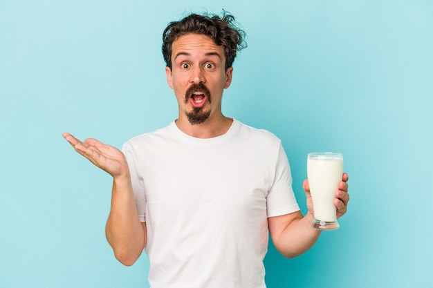 Young caucasian man holding a glass of milk isolated on blue background surprised and shocked.