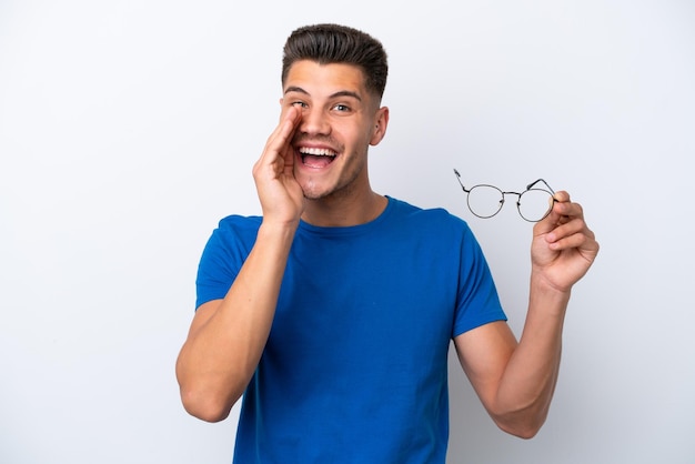 Young caucasian man holding glasses isolated on white background shouting with mouth wide open