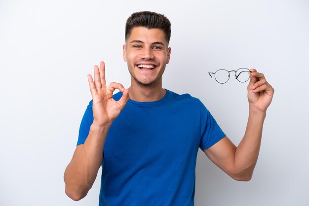 Young caucasian man holding glasses isolated on white background showing ok sign with fingers