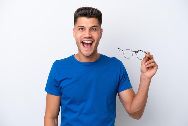 Young caucasian man holding glasses isolated on white background with surprise and shocked facial expression