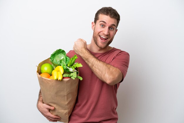 Young caucasian man holding a grocery shopping bag isolated on white background celebrating a victory