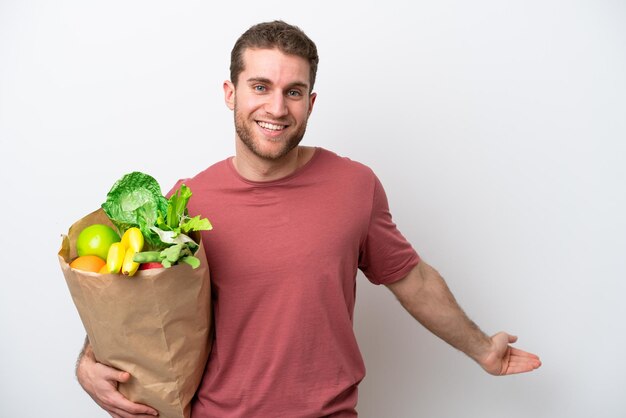 Young caucasian man holding a grocery shopping bag isolated on white background extending hands to the side for inviting to come