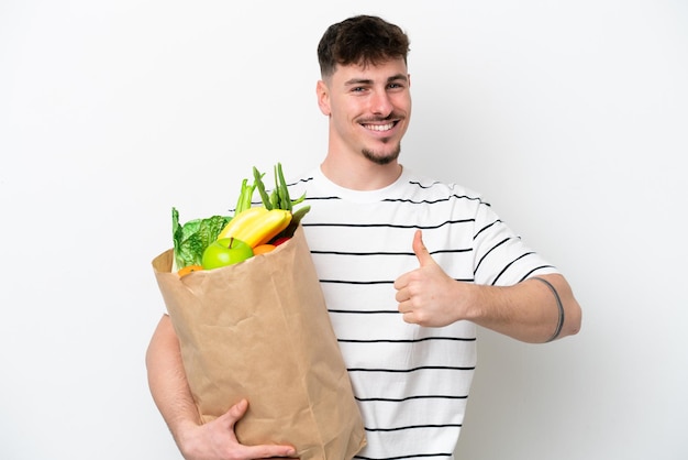 Young caucasian man holding a grocery shopping bag isolated on white background giving a thumbs up gesture