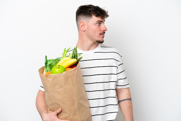 Young caucasian man holding a grocery shopping bag isolated on white background looking to the side