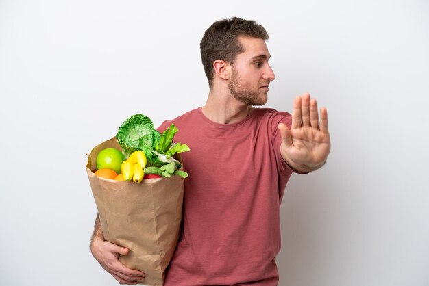 Young caucasian man holding a grocery shopping bag isolated on white background making stop gesture and disappointed