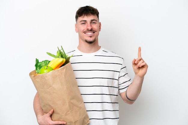 Young caucasian man holding a grocery shopping bag isolated on white background showing and lifting a finger in sign of the best