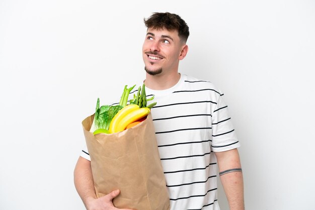 Young caucasian man holding a grocery shopping bag isolated on white background thinking an idea while looking up