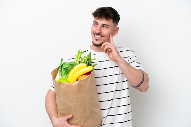 Young caucasian man holding a grocery shopping bag isolated on white background thinking an idea while looking up