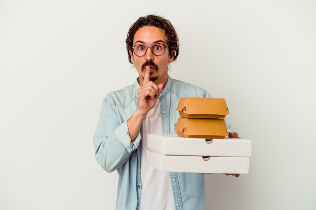 Young caucasian man holding hamburger an pizzas isolated on white keeping a secret or asking for silence.