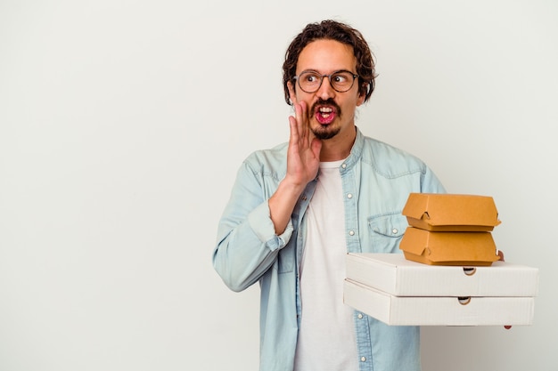 Young caucasian man holding hamburger an pizzas isolated on white wall is saying a secret hot braking news and looking aside