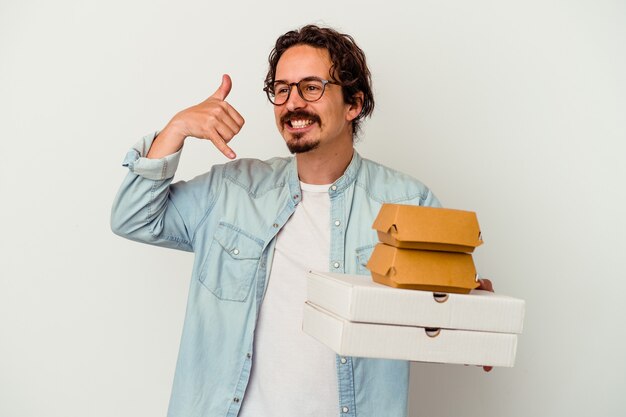 young caucasian man holding hamburger an pizzas isolated on white wall showing a mobile phone call gesture with fingers.