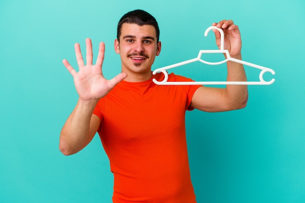 Young caucasian man holding a hanger on blue smiling cheerful showing number five with fingers.