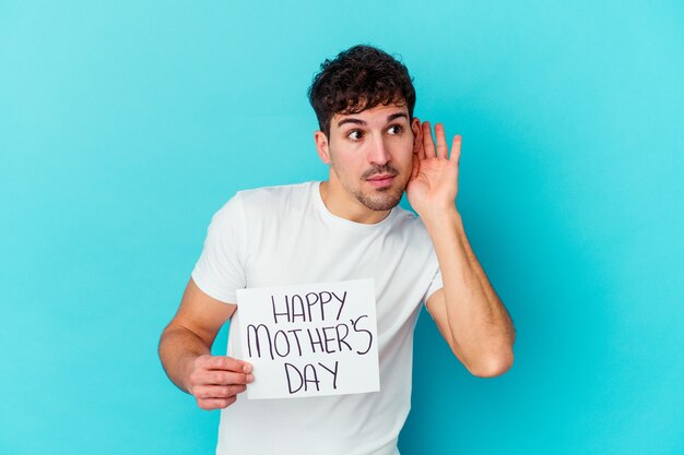 Young caucasian man holding a happy mothers day placard isolated trying to listening a gossip.