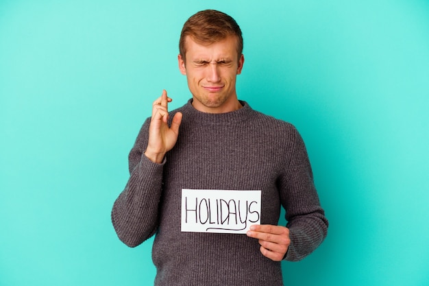 Young caucasian man holding a Holidays placard isolated on blue background crossing fingers for having luck