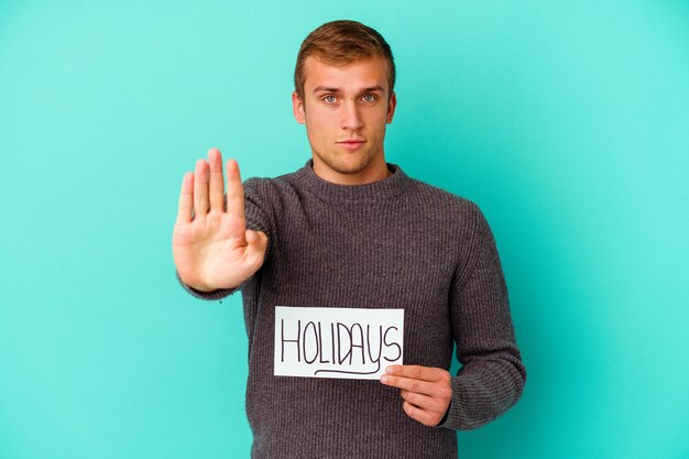 Молодой кавказский человек держит плакат праздников, изолированные на синей стене, стоящей с протянутой рукой, показывая знак остановки, предотвращая вас.