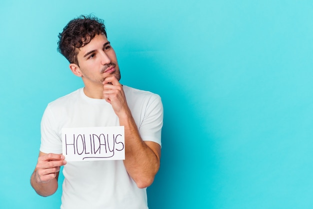 Young caucasian man holding a holidays placard isolated looking sideways with doubtful and skeptical expression.