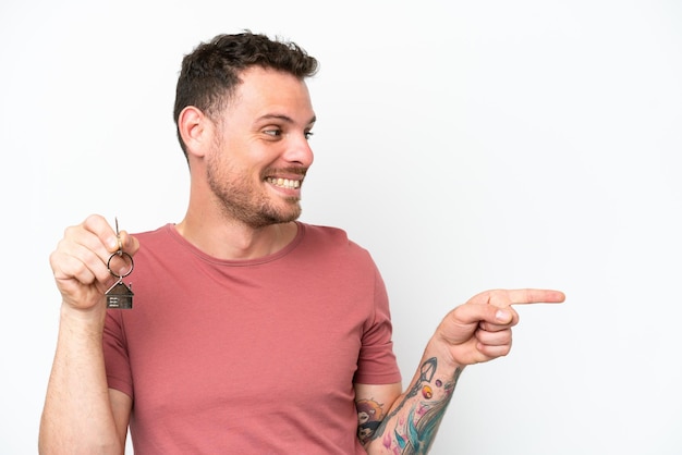 Young caucasian man holding home keys isolated on white background pointing to the side to present a product