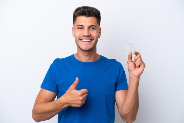 Young caucasian man holding invisible braces isolated on white background with thumbs up because something good has happened