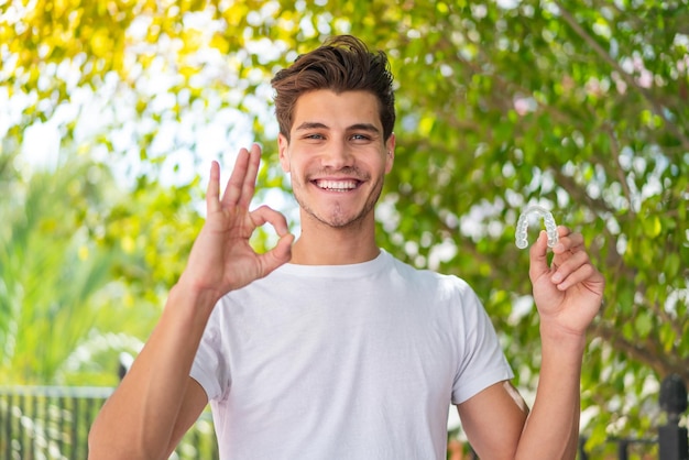 Foto giovane uomo caucasico che tiene bretelle invisibili all'aperto mostrando segno ok con le dita