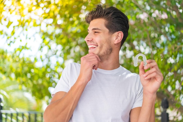 Young caucasian man holding invisible braces at outdoors thinking an idea and looking side