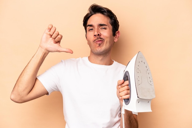 Young caucasian man holding an iron isolated on beige background feels proud and self confident, example to follow.