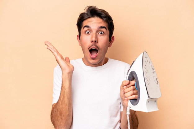 Young caucasian man holding an iron isolated on beige background surprised and shocked