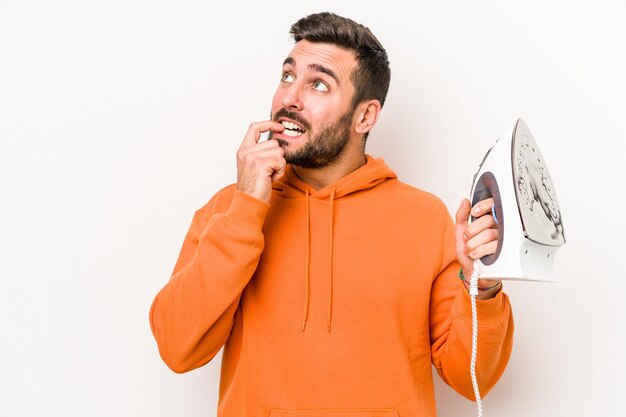 Young caucasian man holding an iron isolated on white background relaxed thinking about something looking at a copy space